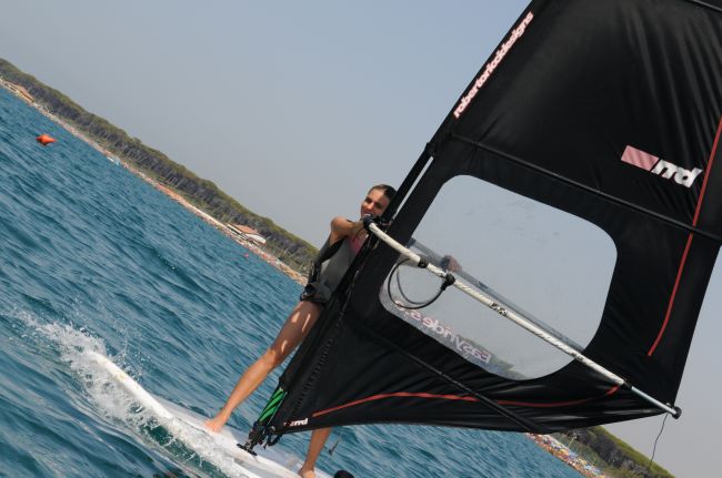 Dettaglio di persona in windsurf vicino alla riva con mare blu e spiaggia sullo sfondo.