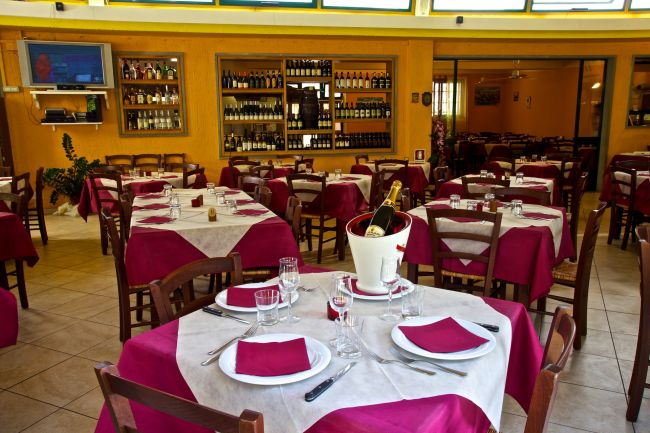 Ristorante con tavoli apparecchiati, tovaglie bordeaux e bottiglie in esposizione.
