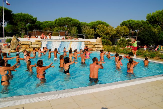 Persone che fanno acquagym in piscina all'aperto, con alberi sullo sfondo.