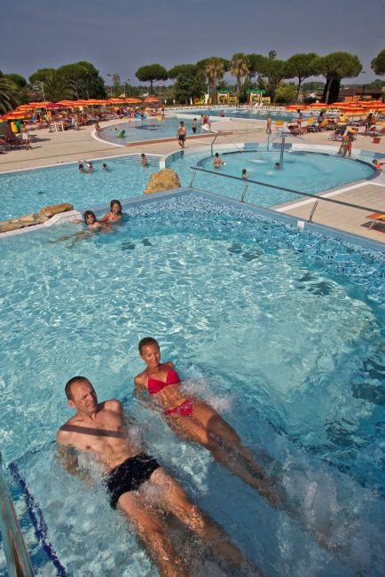 Una piscina con persone che si rilassano, sedie a sdraio e ombrelloni in fondo.