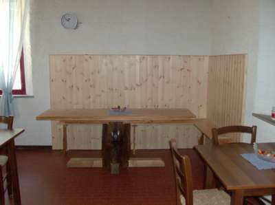 Un tavolo di legno con sedie in una stanza semplice, pareti bianche e una mensola di legno chiaro.
