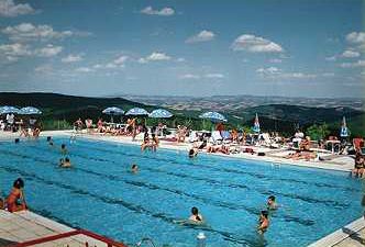 Piscina all'aperto con bagnanti e lettini, colline sullo sfondo.