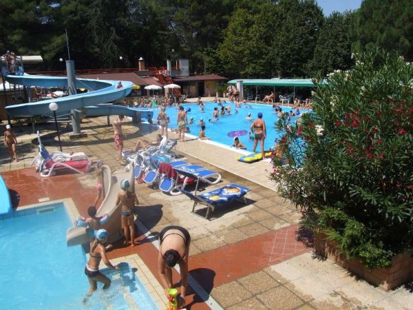 Piscina affollata con scivoli e lettini. Alberi sullo sfondo.