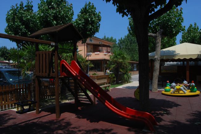 Parco giochi con scivolo rosso, altalene e giochi a molla. Alberi e recinzione sullo sfondo.