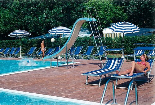 Piscina con sdrai, ombrelloni a righe e uno scivolo. Un bagnante prende il sole.