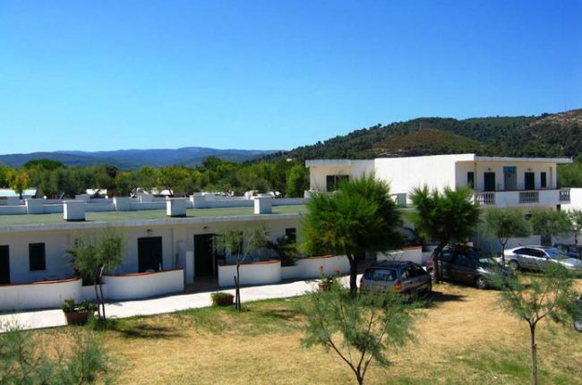 Edificio bianco con parcheggio e alberi, colline sullo sfondo.