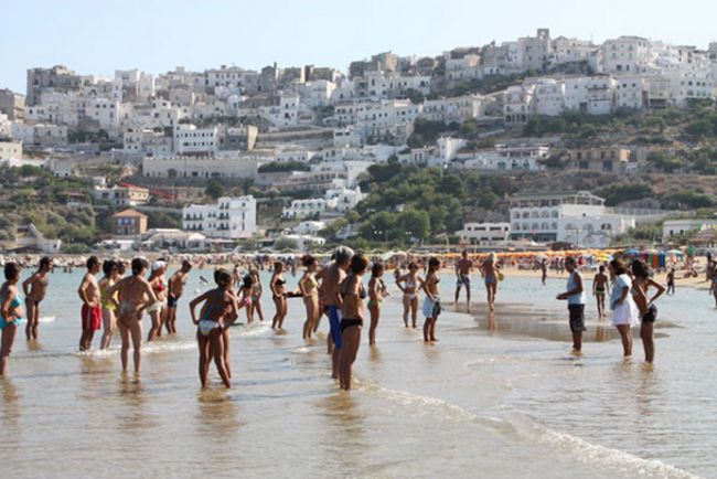 Persone in acqua su una spiaggia, case bianche sulla collina.