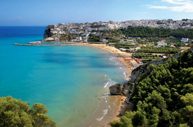 Spiaggia panoramica con mare azzurro e città collinare sullo sfondo.